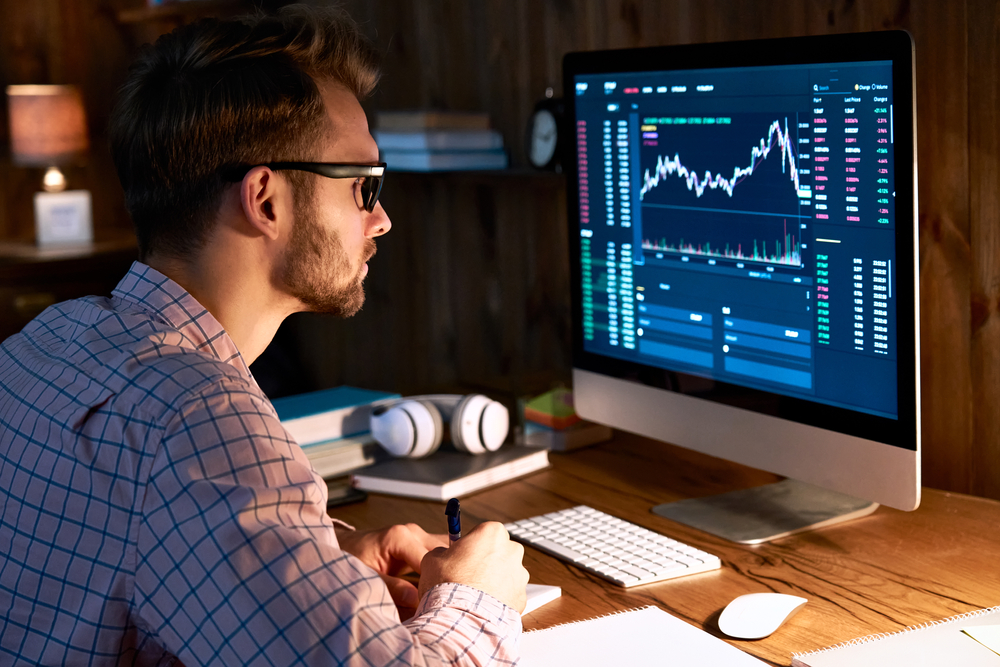 Man with glasses writing notes as he monitors a computer screen showing stock charts and data.