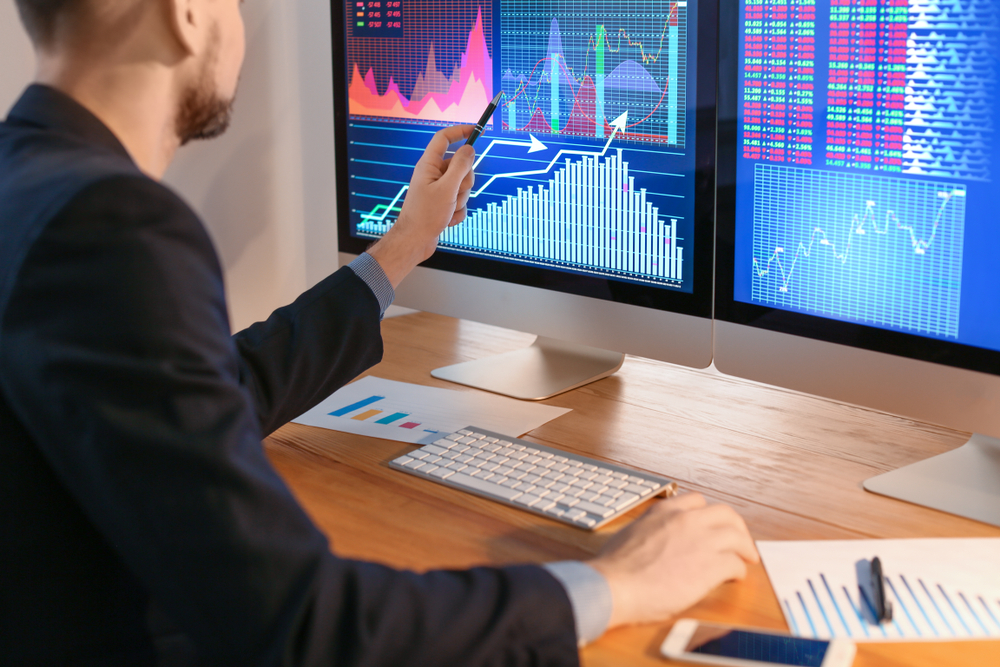 Man in a suit analyzes stock charts on dual monitors, pointing with a pen at the data.