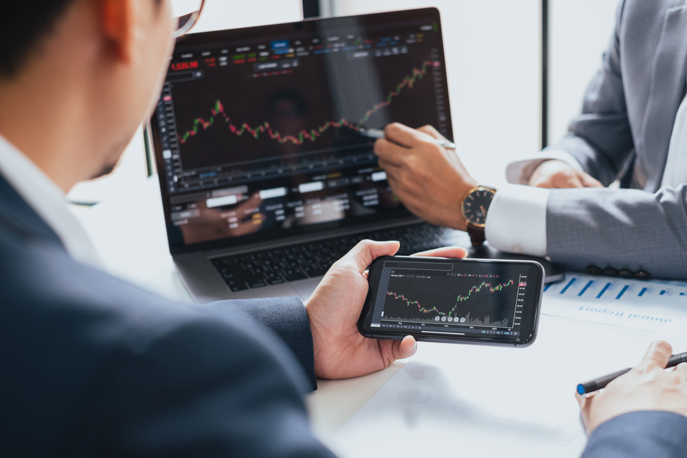 Two professionals in suits review stock charts on a laptop and a smartphone during a meeting.