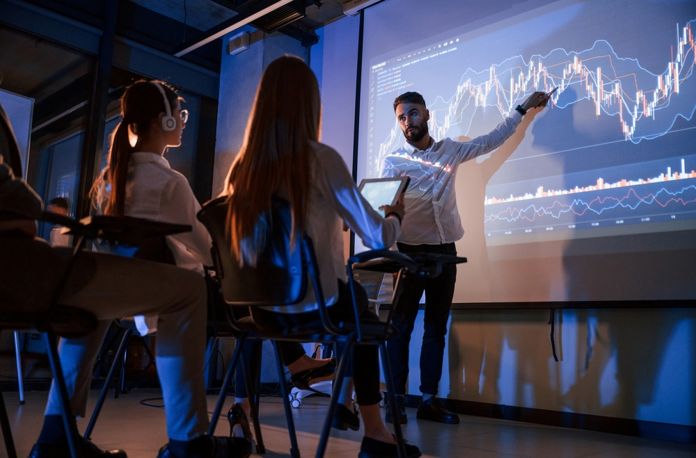 Man leads a finance class, pointing to a large stock-chart projection on a screen while students watch with tablets and notebooks in a dark classroom.