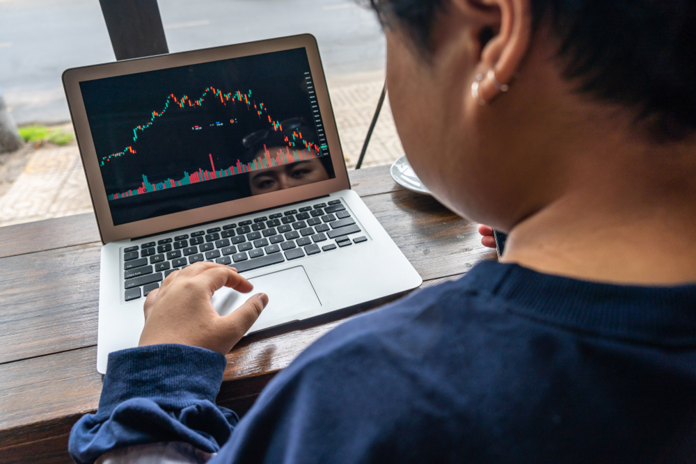 Person using a laptop at a wooden table, screen showing a stock market chart with green and red candlesticks and volume bars.